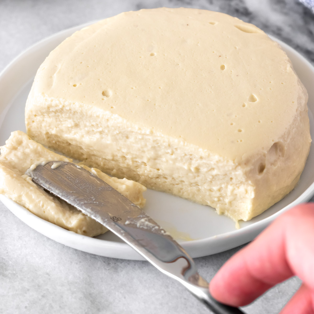 Queijo de castanha-de-caju redondo em cima de um prato. Uma faca está fatiando um lado do queijo.