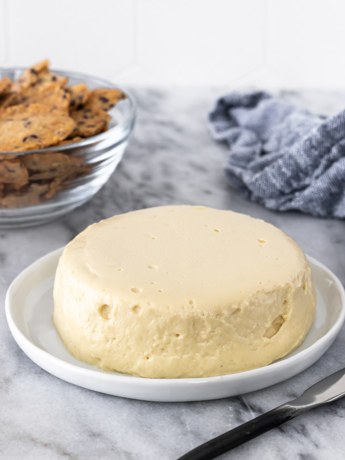 Queijo de castanha-de-caju redondo em cima de um prato. Ao lado dele, biscoitos de sementes estão servidos em um pote para acompanhar o queijo.