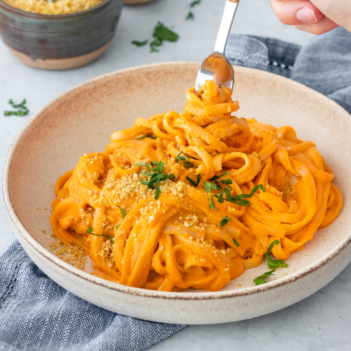 garfo sendo retirado de prato de macarrão envolvido com molho de pimentão vermelho assado, servido com parmesão vegano de castanhas e salsinha fresca picada